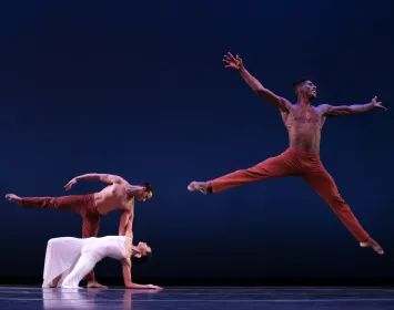Tres bailarines danzan , uno de ellos salta y tiene los brazos abiertos.