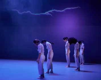 Cinco bailarinas danzan, tienen su cabezas mirando hacía el suelo. Un rayo esta proyectado en la parte de arriba. 