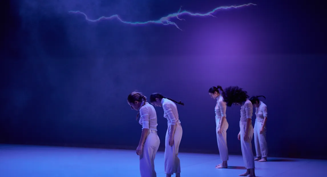 Cinco bailarinas danzan, tienen su cabezas mirando hacía el suelo. Un rayo esta proyectado en la parte de arriba.