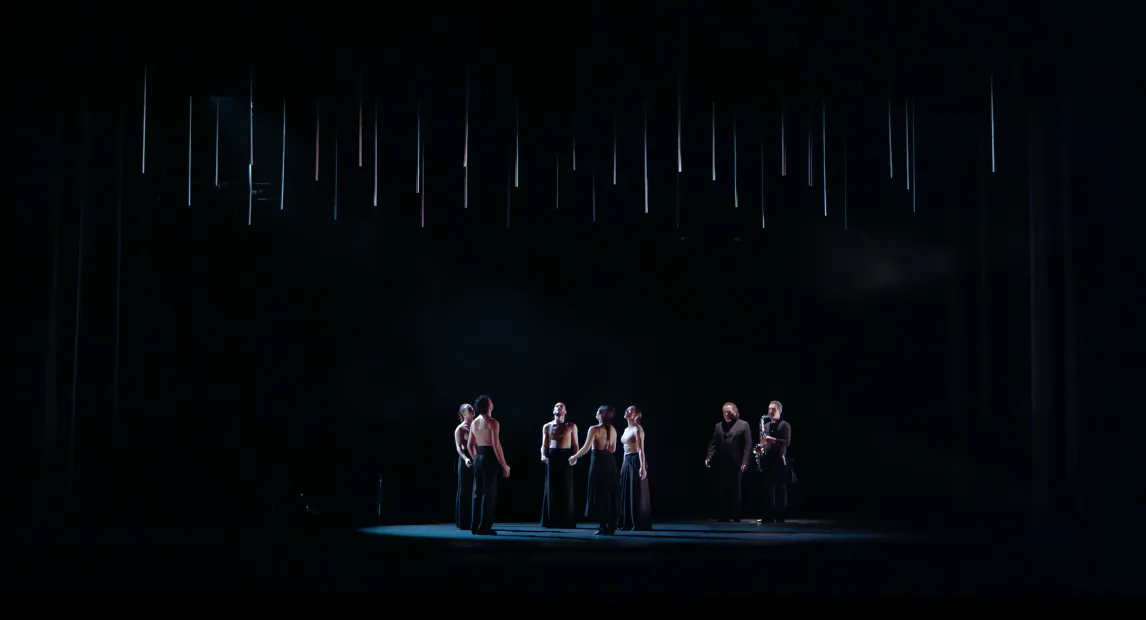 Un grupo de bailarines miran arriba, las luces bajan.