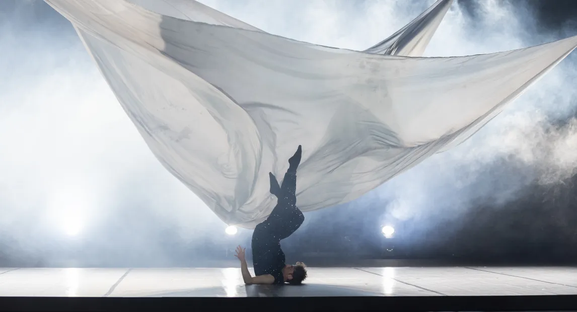 Un bailarín danza, tiene los pies hacía arriba y su cabeza sobre el suelo, hay una gran sabana blanca detrás de él haciendo figuras.