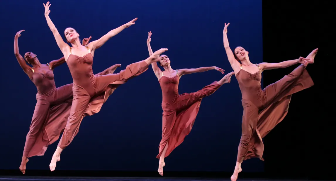 Cuatro bailarines danzan, tienen vestidos largos, sus piernas y brazos los tienen levantados.