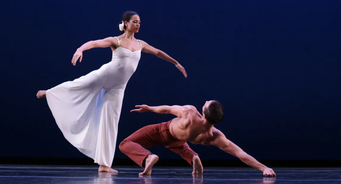 Dos bailarines danzan, una mujer esta de pie subiendo los brazos y un hombre esta sobre el suelo.