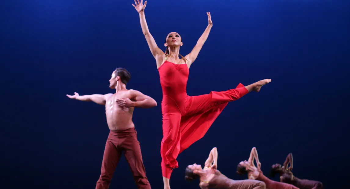 un grupo de bailarines danzan. Una mujer salta , sus pies no tocan el suelo y sus brazos están extendidos