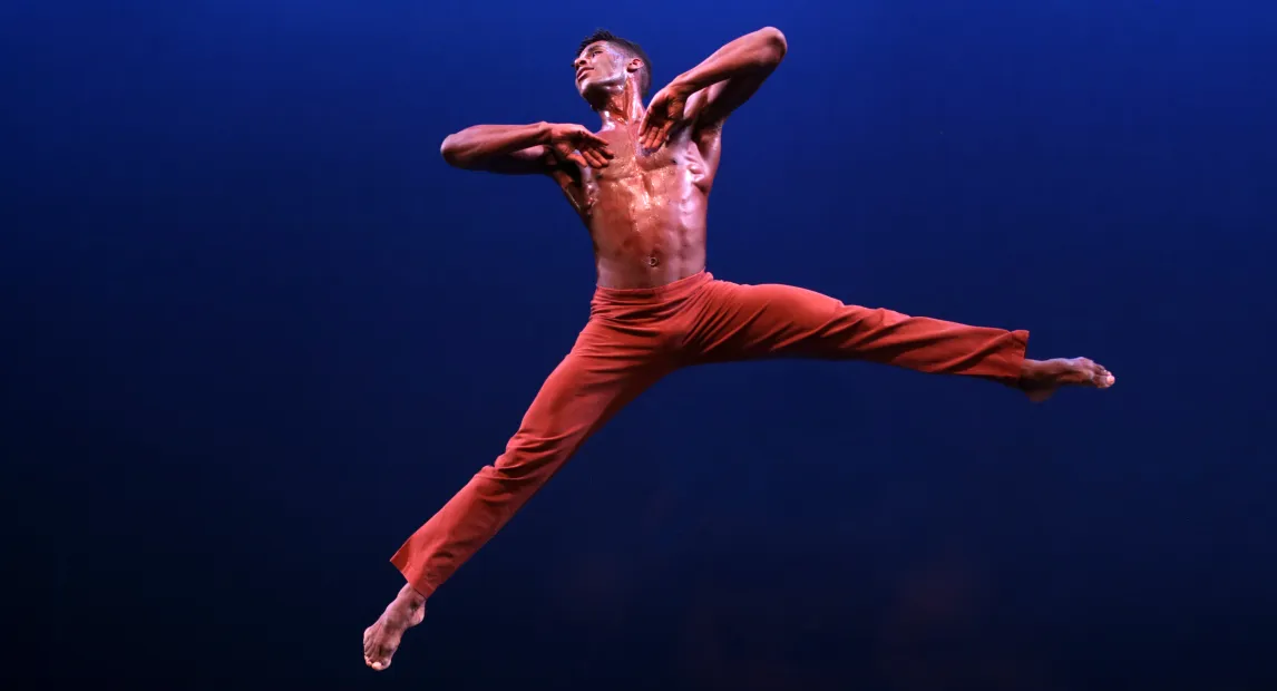 Un bailarín está sostenido en el aire. tiene un pantalón rojo. Sus pies se estiran en el aire.