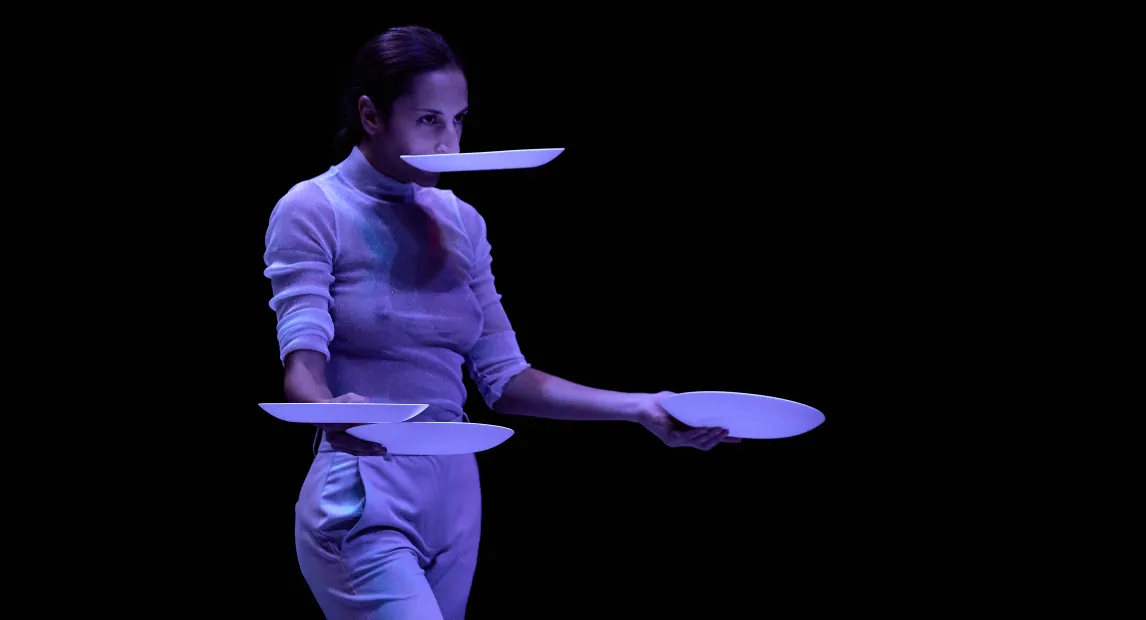Una bailarina sostiene un plato en su boca y tres platos en sus manos.