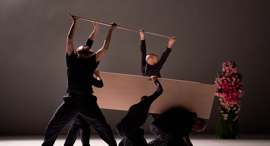 Un grupo de bailarines danzan, sujetan una madera entre ellos como parte del performance.