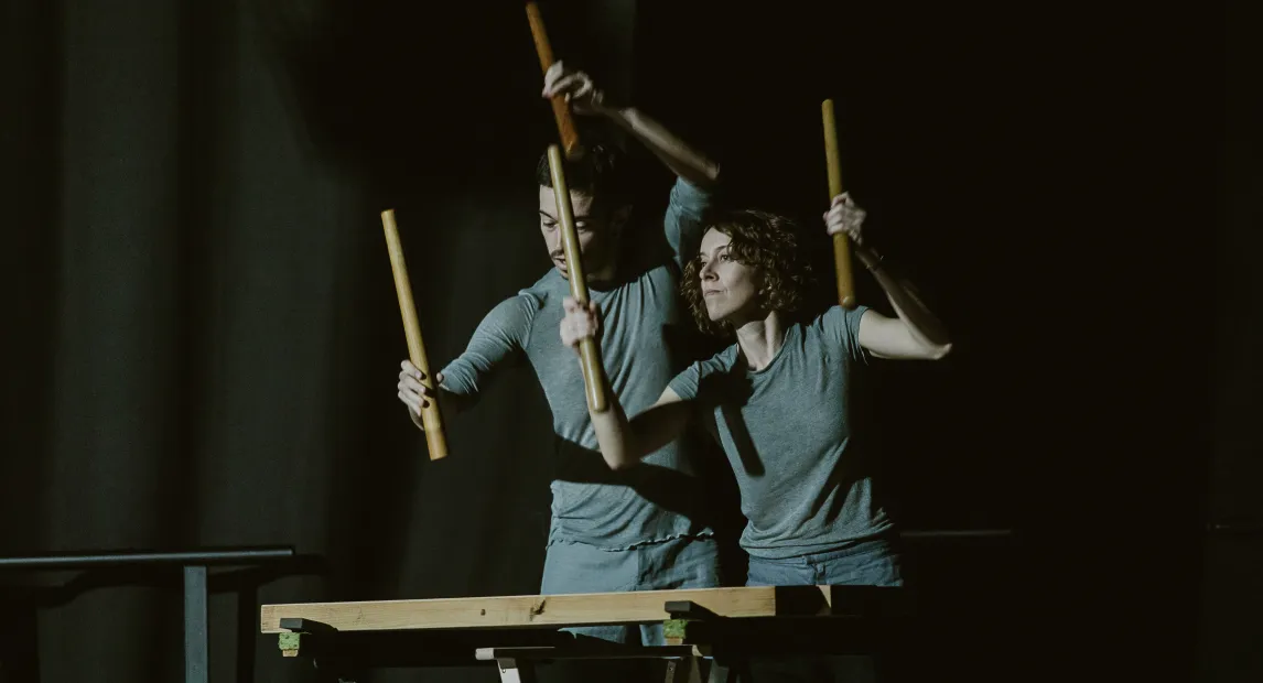 Dos bailarines tocan un instrumento, tienen en sus manos palos de madera con los que producen el sonido
