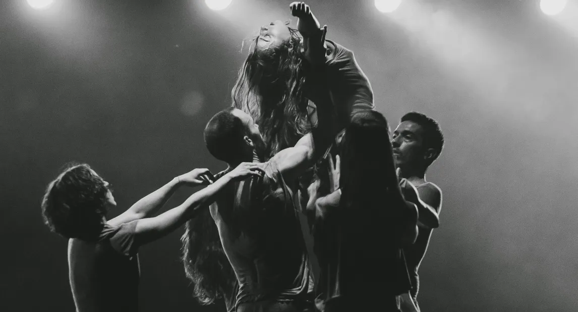 Un grupo de bailarines danzan y tienen alzada a una del grupo que a su vez tiene los brazos abiertos.