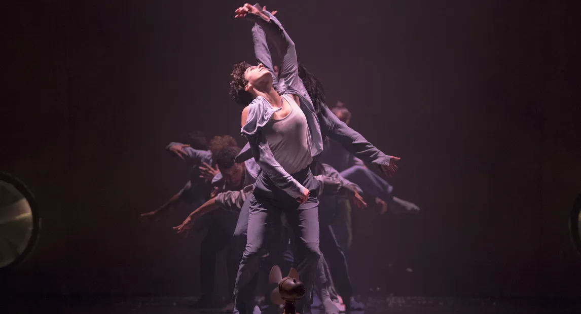 Un grupo de bailarines danzan, el bailarín principal tiene la mano alzada.