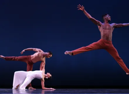 Tres bailarines danzan , uno de ellos salta y tiene los brazos abiertos.