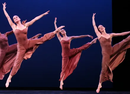 Cuatro bailarines danzan, tienen vestidos largos, sus piernas y brazos los tienen levantados.
