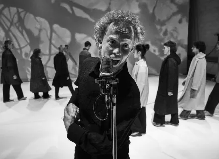 Hombre cantando en un microfóno de pie en primer término. Detrás una fila de personas vestidas con abrigos andan en fila india