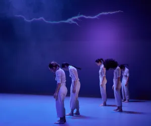 marcos G punto Cinco bailarinas danzan, tienen su cabezas mirando hacía el suelo. Un rayo esta proyectado en la parte de arriba.