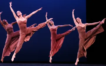 Cuatro bailarines danzan, tienen vestidos largos, sus piernas y brazos los tienen levantados. 