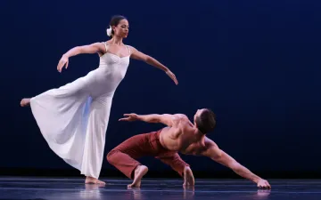 Dos bailarines danzan, una mujer esta de pie subiendo los brazos y un hombre esta sobre el suelo.