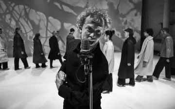 Hombre cantando en un microfóno de pie en primer término. Detrás una fila de personas vestidas con abrigos andan en fila india 